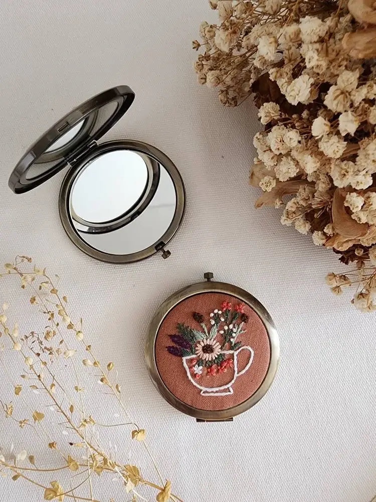 Compact mirror with floral design on a light surface with dried flowers.
