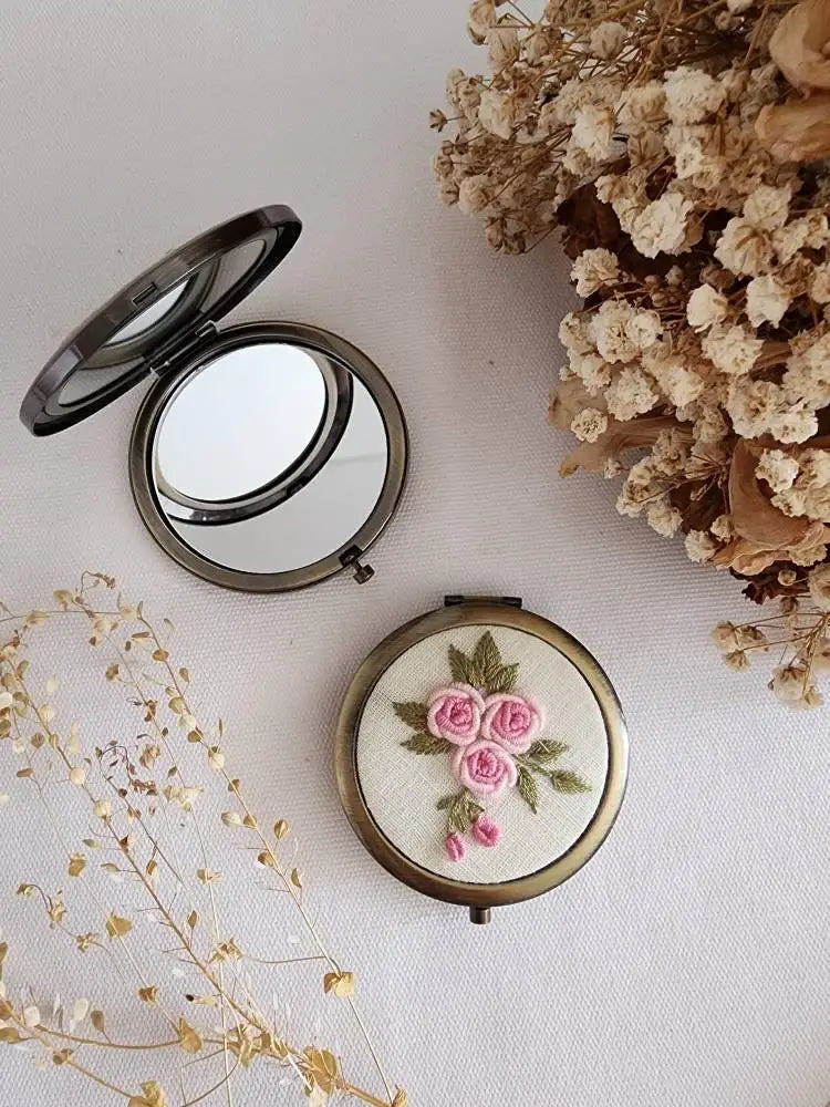Compact mirror with embroidered flowers on a light surface with dried flowers around