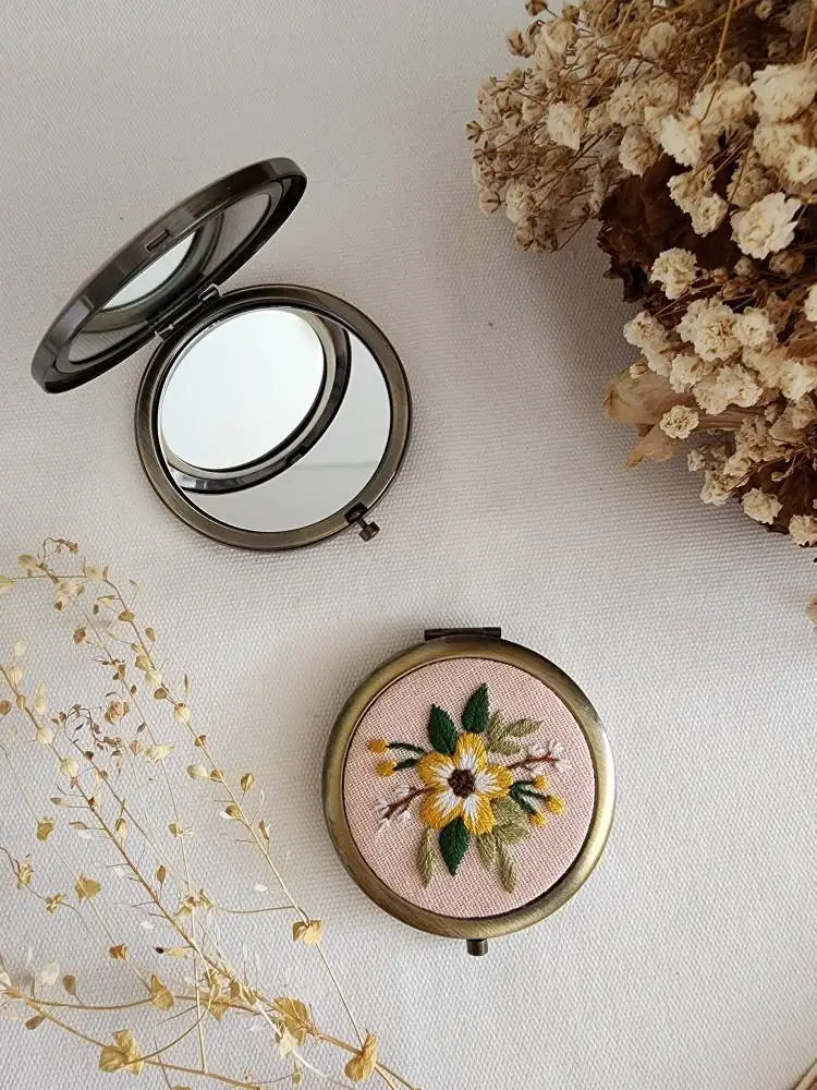 Compact mirror with floral embroidery on a light surface with dried flowers.
